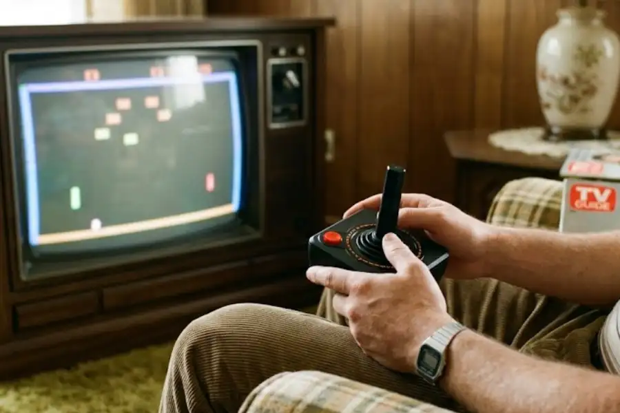 Hands holding an Atari 2600 joystick controller in front of a vintage CRT television displaying a retro game