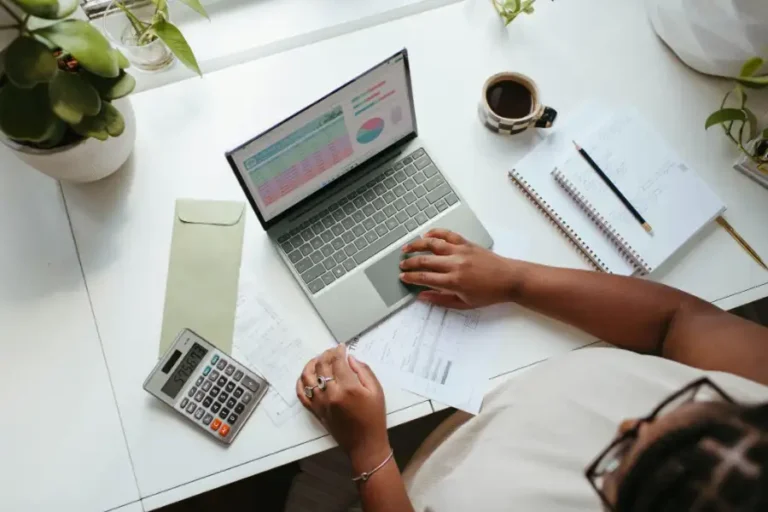 Person working on a laptop at a desk with financial documents, calculator, notebook, and coffee.