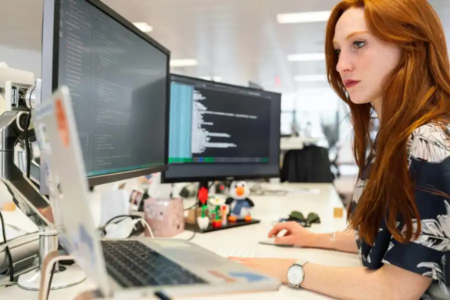 A woman working on computer code across multiple monitors in a modern office.