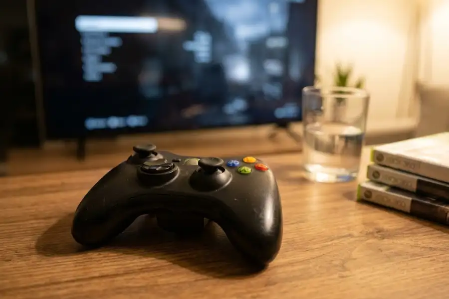 Xbox 360 controller on a desk with a TV showing a cheat activated message in the background
